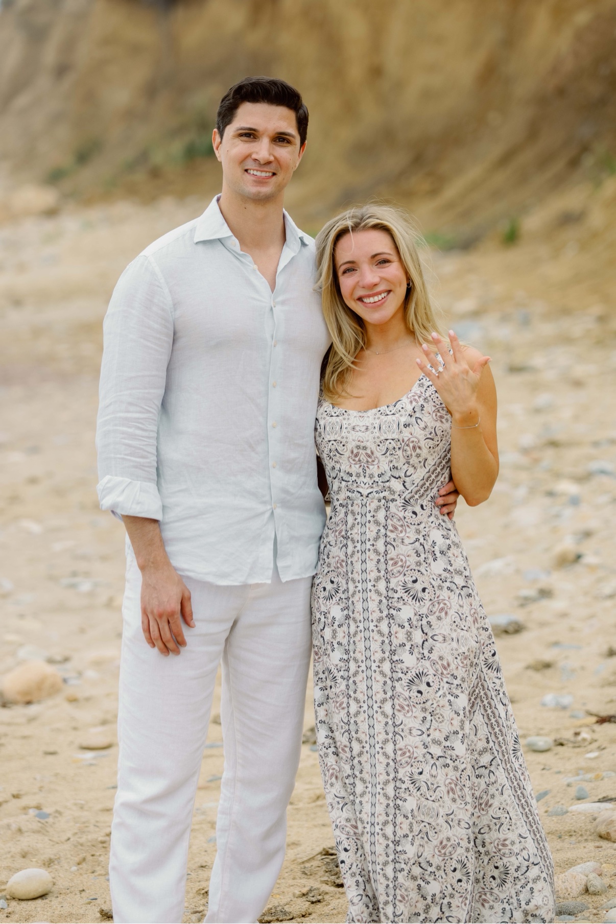 Newly engaged couple on the beach at Montauk Lighthouse, Long Island — she raises her hand to show the ring, the bluff behind them