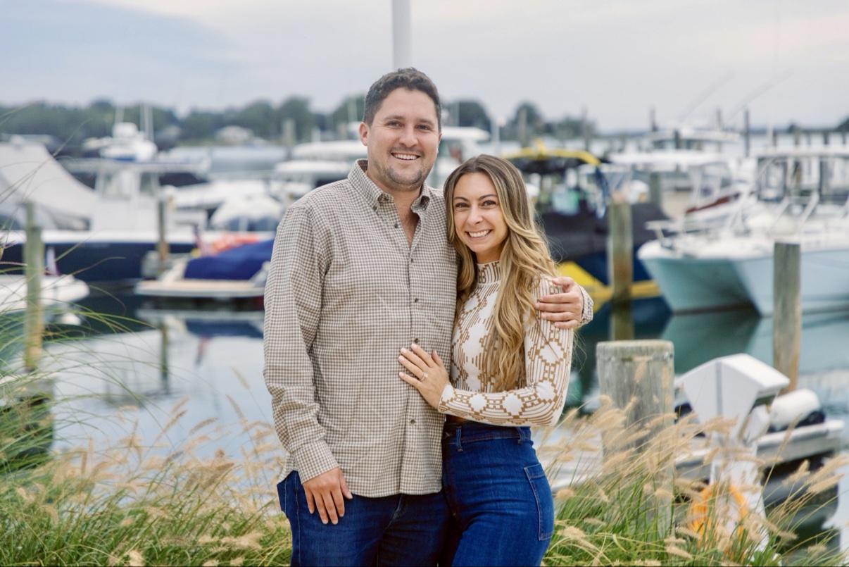 Newly engaged couple standing in the tall grasses at the Sag Harbor marina, Long Island, boats behind them