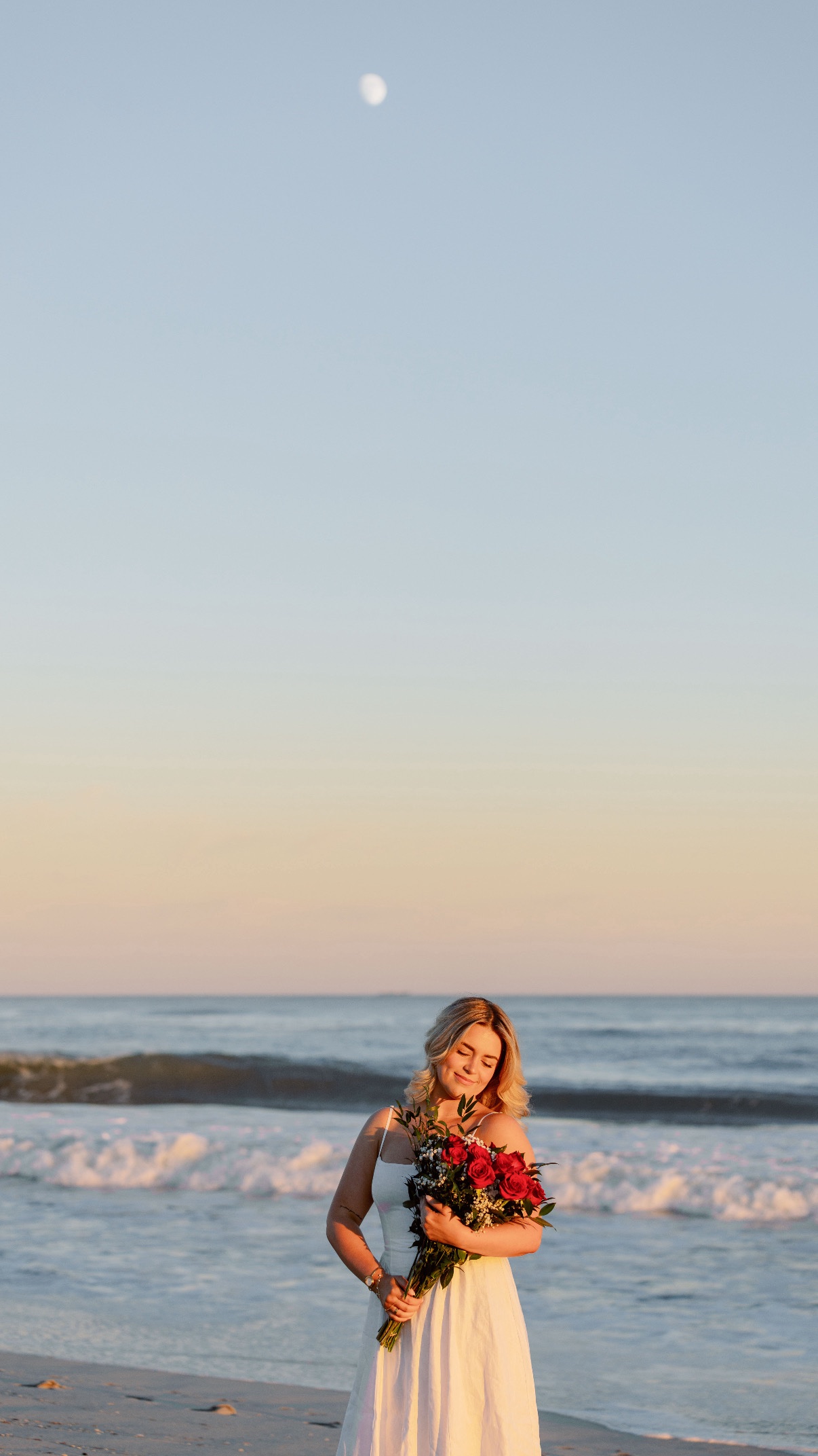 Engagement photos at Cooper's Beach, Southampton, Long Island — the bride-to-be alone with her red rose bouquet at the water's edge, the moon already up in the evening sky