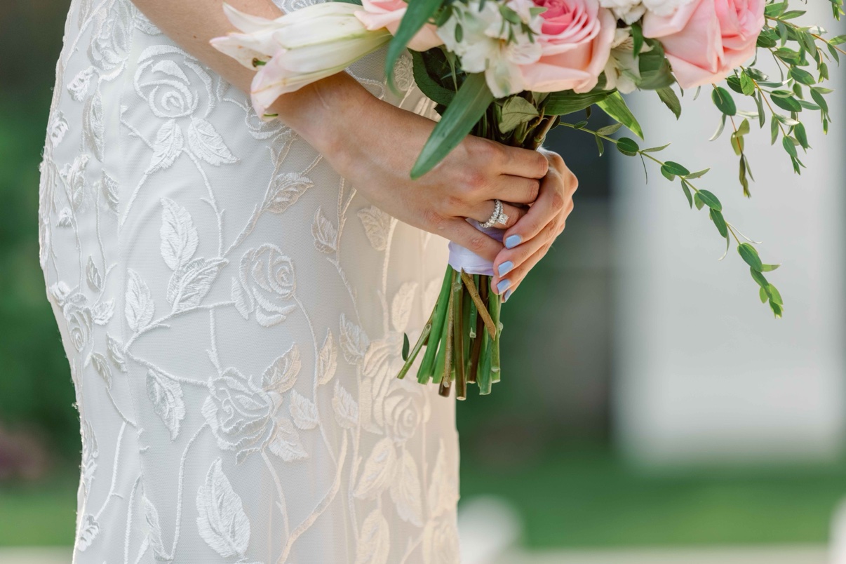 Close-up of the bride’s hands holding her bouquet over her lace gown, her ring visible