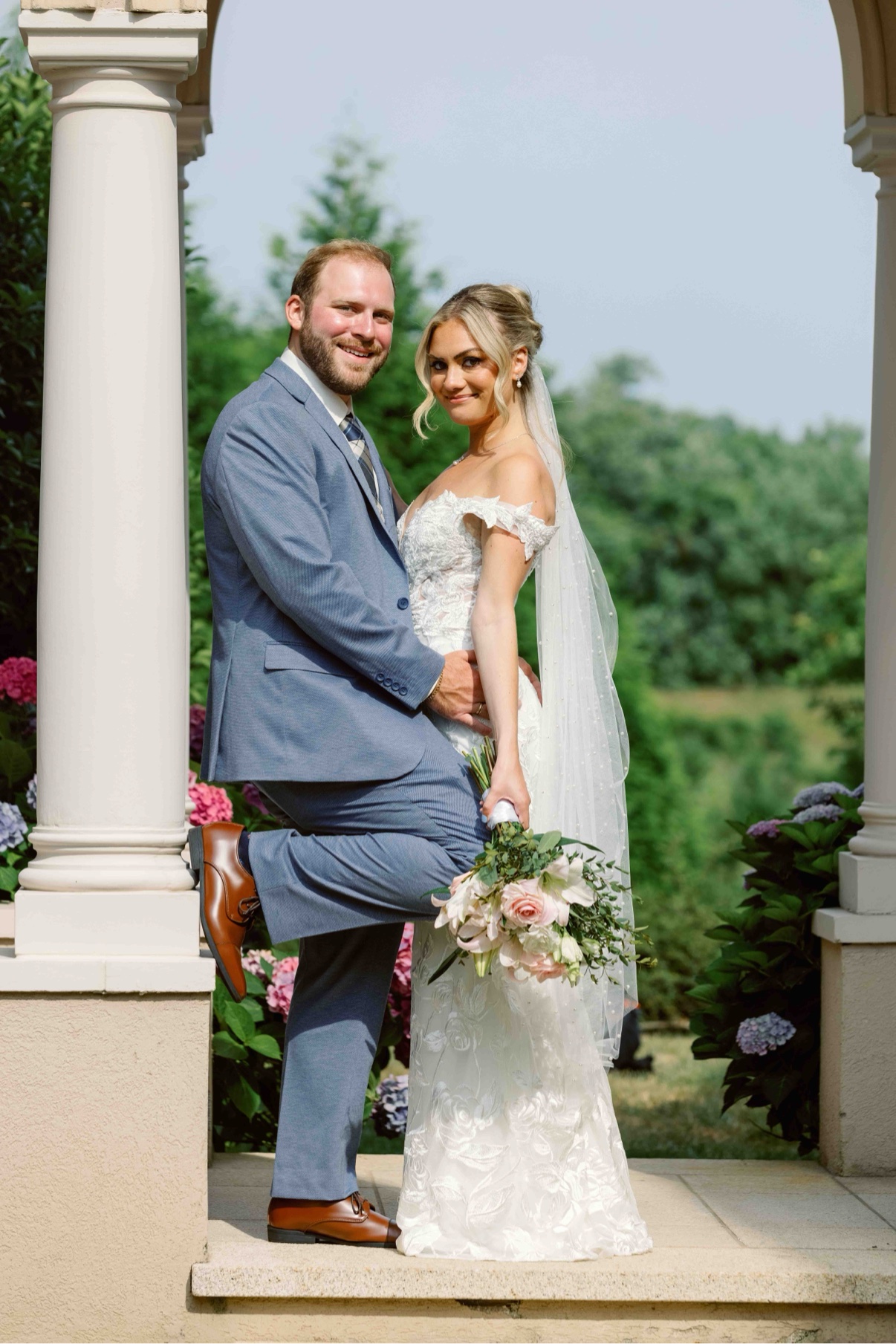Bride and groom posed beside white columns with her veil flowing