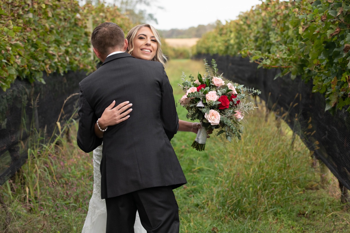 Bride smiling over the groom’s shoulder as they embrace between vineyard rows